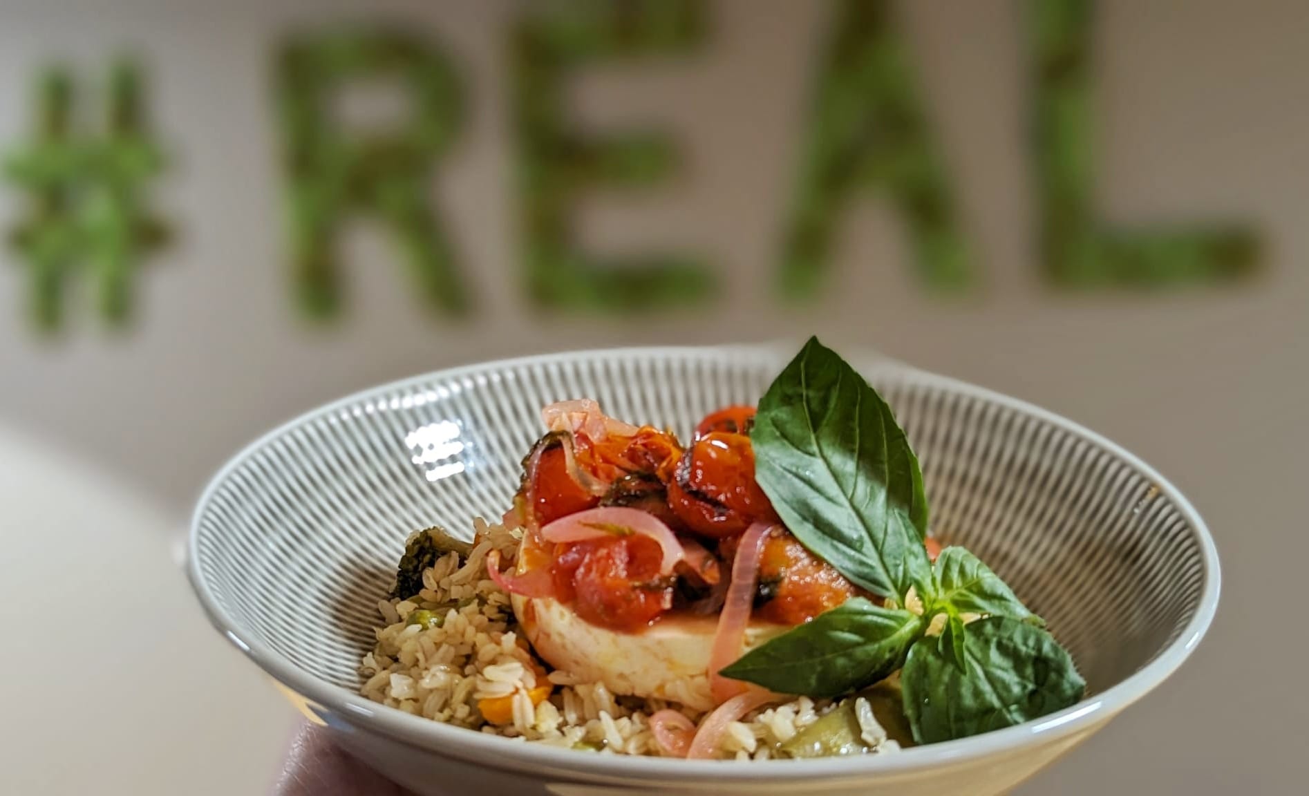 Bowl mit Fisch, Tomaten und Basilikum vor dem #REAL-Schriftzug im Betriebsrestaurant