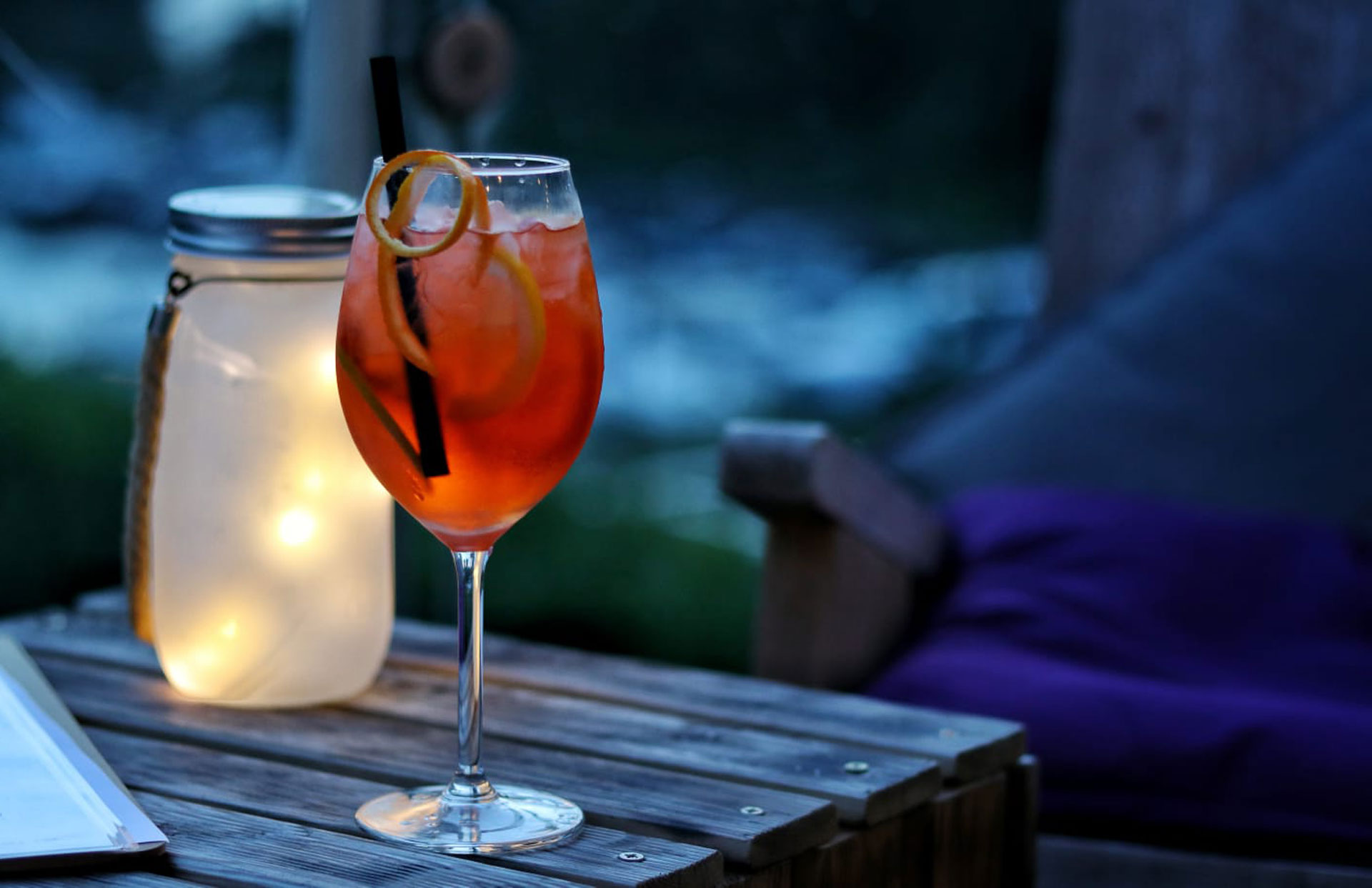 Getränk mit Orangenscheibe im Weinglas neben einem Windlicht auf einem Holztisch am Abend